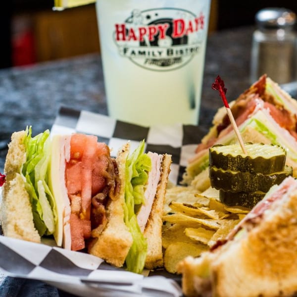 Happy Days Finer Diner club sandwich with chips, pickle, and a drink with the restaurant logo.