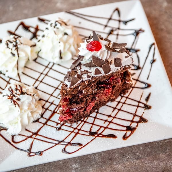 A slice of Black Forest cake with whipped cream, chocolate shavings, and a cherry on a white plate.