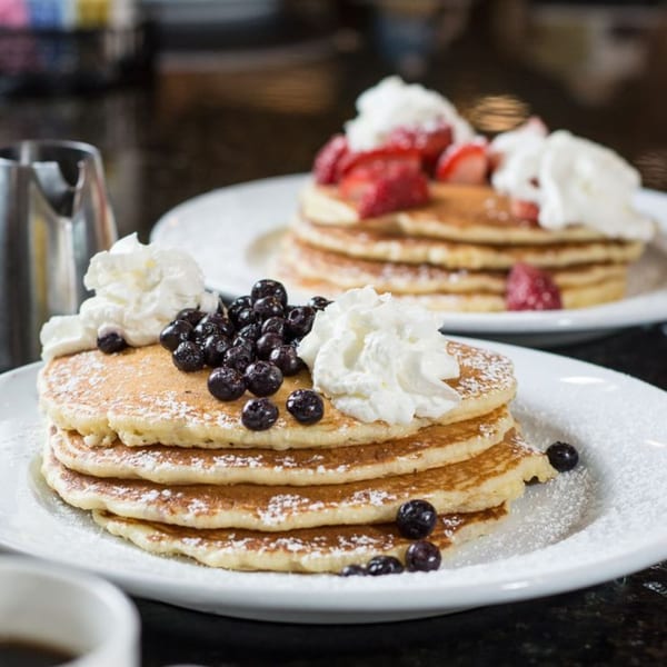 Two plates of diner pancakes, one with blueberries and whipped cream, the other with strawberries.